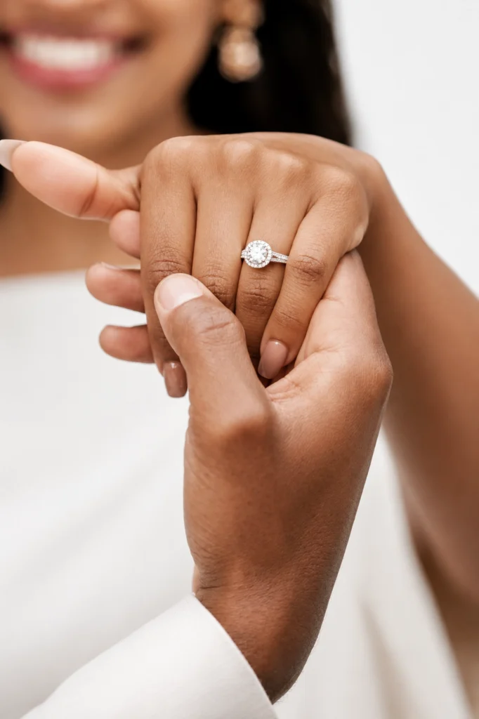 A close-up of two hands, with one hand wearing a stunning diamond engagement ring. The person wearing the ring is smiling in the background, perfectly capturing the joy and elegance that Diamond Engagement Rings symbolize.